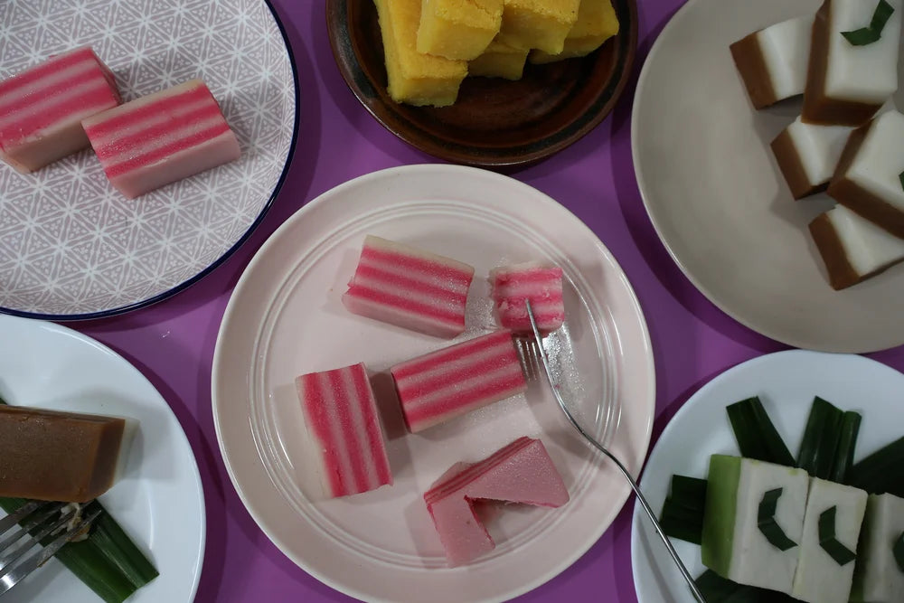 Assorted desserts including pink and white layered cake, yellow sponge cake, and green and white layered cake on a purple table.