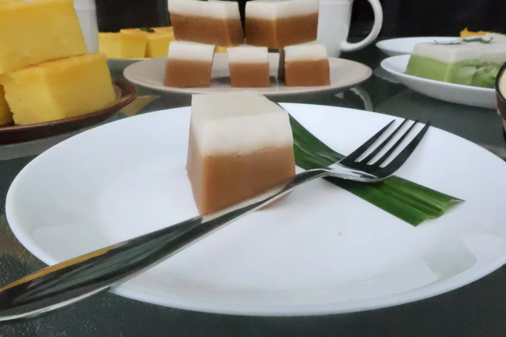 Square piece of cake on a white plate with a fork, surrounded by other desserts.